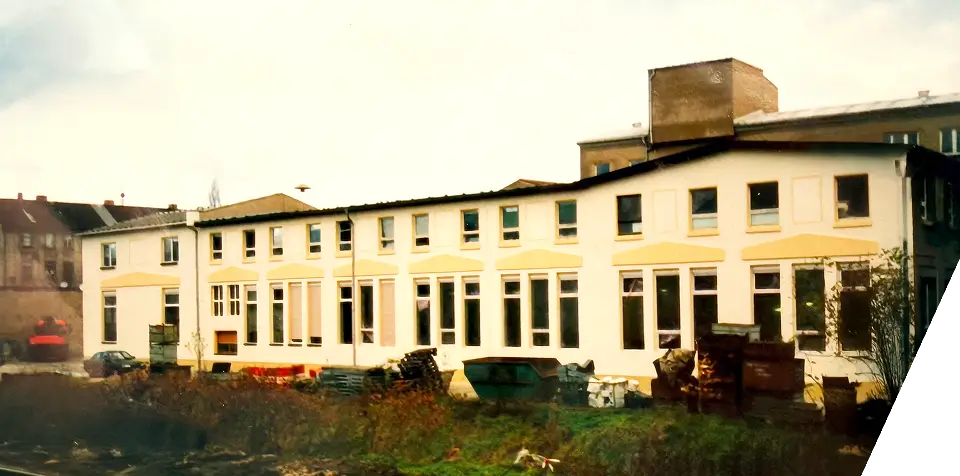 Ein zweistöckiges weißes Gebäude mit gelben Zierleisten steht hinter überwucherndem Gras und Schutthaufen. Das Gebäude hat mehrere Fenster, und im Hintergrund ist ein größerer Backsteinbau zu sehen.