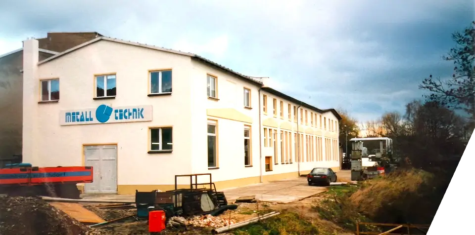 Ein helles Industriegebäude mit der Aufschrift Metall Technik, mehrere geparkte Fahrzeuge, Baumaterialien und ein bewölkter Himmel im Hintergrund. In der Nähe des Gebäudes sind Bäume und Gras zu sehen.