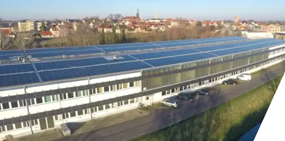 Ein langgestrecktes, weißes Industriegebäude mit zahlreichen Solarzellen auf dem Dach, davor mehrere geparkte Autos und im Hintergrund eine entfernte Stadt mit verschiedenen Gebäuden und einem Kirchturm.