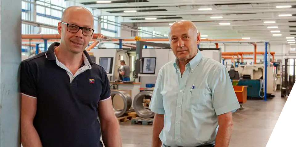 Zwei Männer stehen in einer modernen Industrieanlage mit Maschinen und Geräten im Hintergrund. Beide tragen Brillen und Freizeithemden, und die Umgebung scheint eine Fabrik oder ein Produktionsbetrieb zu sein.