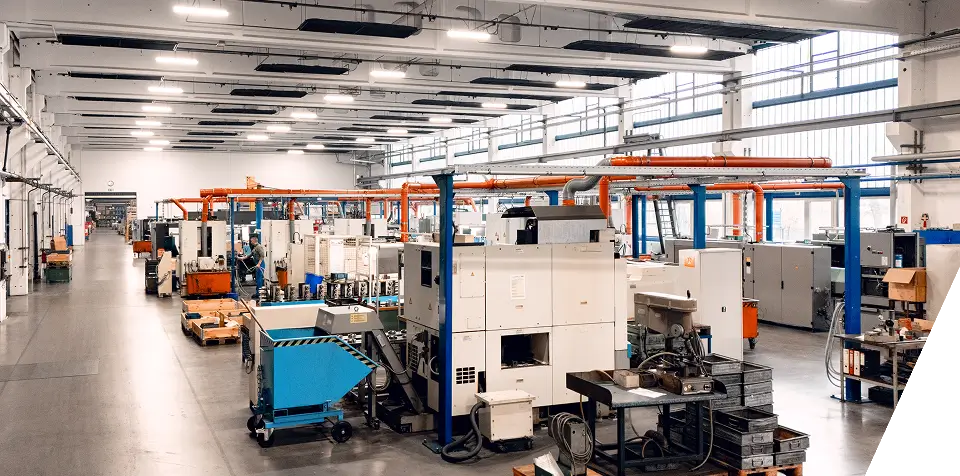 Große, saubere Fabrikhalle mit Reihen von Industriemaschinen, Arbeitsplätzen und orangefarbenen Deckenrohren. Der Raum ist gut beleuchtet, hat hohe Decken, breite Gänge und geordnete Geräte. Es sind keine Arbeiter zu sehen.