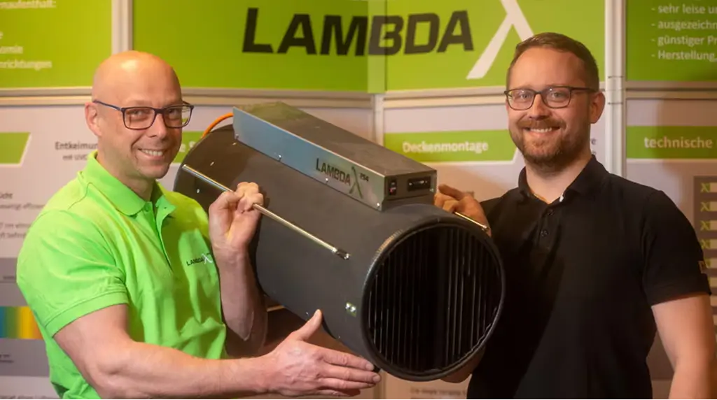 Zwei lächelnde Männer halten ein großes, zylindrisches schwarzes Gerät mit der Aufschrift LAMBDA vor einem Ausstellungsstand mit dem LAMBDA-Logo und technischen Informationen.