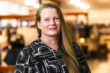 Eine Frau mit langen blonden Haaren und einer schwarz-weiß gemusterten Bluse steht in einem Haus und lächelt sanft, während der Hintergrund mit hellen Lichtern und Industrieanlagen verschwimmt.