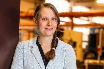 Eine Frau mit hellbraunem, zu einem Zopf geflochtenem Haar, die einen hellblauen, strukturierten Blazer über einem dunklen Hemd trägt, lächelt, während sie in einer Industrie- oder Laborumgebung mit verschwommenen Geräten im Hintergrund steht.