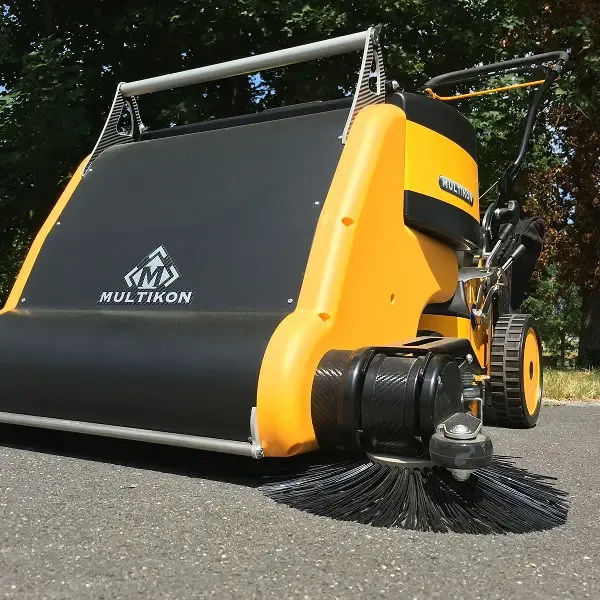 Nahaufnahme einer gelb-schwarzen Multikon-Kehrmaschine auf einer asphaltierten Straße, wobei der vordere Bürstenaufsatz sichtbar ist und Bäume im Hintergrund zu sehen sind.