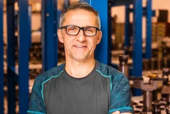 Ein Mann mittleren Alters mit kurzen grauen Haaren und Brille steht lächelnd mit verschränkten Armen da und trägt ein graues Sporthemd. Hinter ihm sind blaue Metallkonstruktionen und Industrieanlagen in einer Werkstatt oder Fabrik zu sehen.