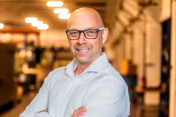 Ein glatzköpfiger Mann mit Brille und weißem Hemd steht lächelnd mit verschränkten Armen in einer hell erleuchteten Industrie- oder Büroumgebung, mit unscharfen Regalen und Lichtern im Hintergrund.