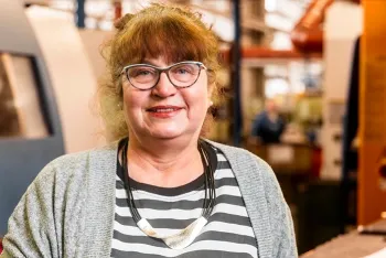 Eine Frau mit Brille, lockigem braunem Haar und gestreiftem Hemd lächelt in einer Industrie- oder Werkstatteinrichtung, trägt eine graue Strickjacke und eine Statement-Halskette.