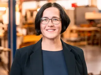 Eine Frau mit kurzen dunklen Haaren, Brille und schwarzem Blazer lächelt, während sie in einer hell erleuchteten Industrie- oder Werkstattumgebung steht.