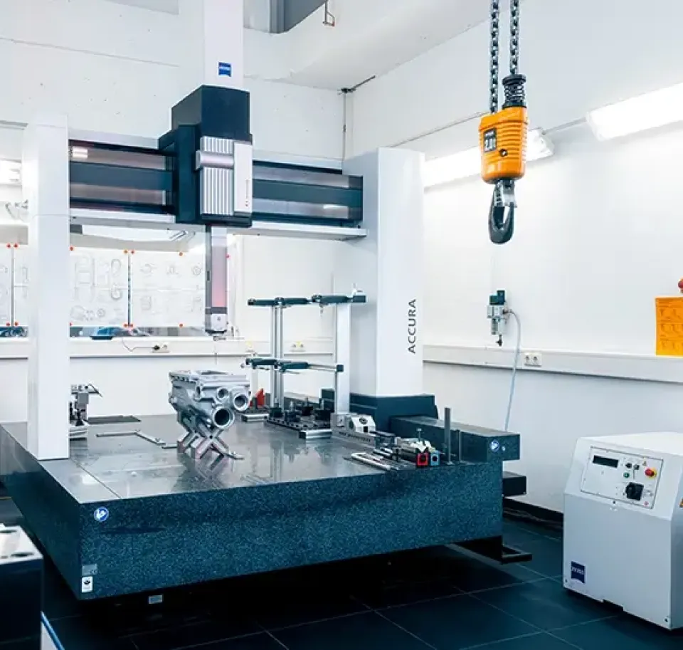 Ein modernes Labor mit einer großen Koordinatenmessmaschine (KMG), einem Metallteil auf dem Messbett, einem hängenden gelben Aufzug, technischen Zeichnungen an der Wand und verschiedenen Geräten in einem sauberen, gut beleuchteten Raum.