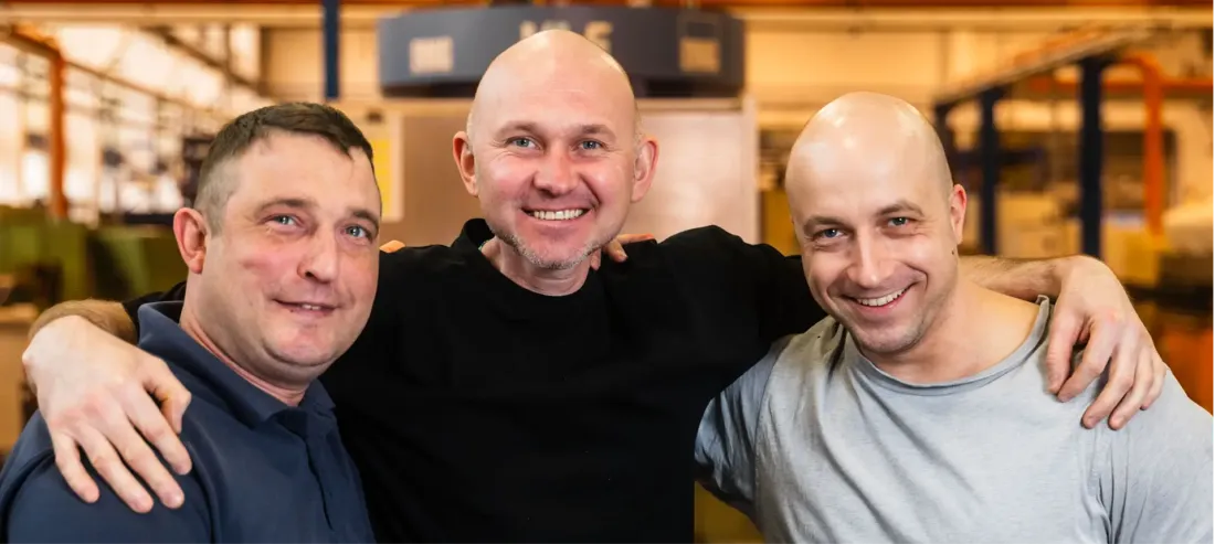 Drei lächelnde Männer, einer in einem marineblauen, einer in einem schwarzen und einer in einem grauen Hemd, stehen in einer Industrie- oder Fabrikumgebung eng beieinander und legen die Arme umeinander.