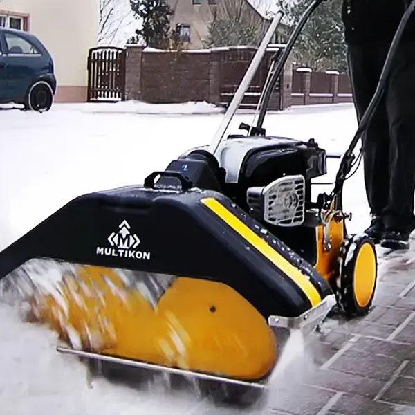 Eine Person benutzt eine Schneeräummaschine der Marke Multikon mit gelben Walzen, um einen gepflasterten Gehweg in einem Wohngebiet vom Schnee zu befreien. Der Boden ist mit Schnee bedeckt und ein Auto ist in der Nähe geparkt.