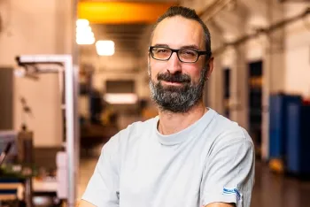Ein bärtiger Mann mit Brille, der ein hellgraues T-Shirt trägt, steht mit verschränkten Armen und lächelt in einer gut beleuchteten Industrie- oder Werkstattumgebung mit unscharfen Maschinen im Hintergrund.