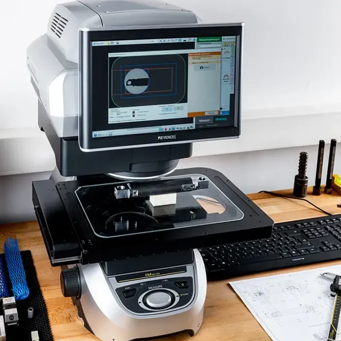 Ein digitales Messmikroskop mit einem Monitor zeigt das Bild eines Objekts und die Messungen an. Das Gerät steht auf einem Holztisch mit einer Tastatur, einem Messschieber, blauen Kabeln und technischen Zeichnungen in der Nähe.