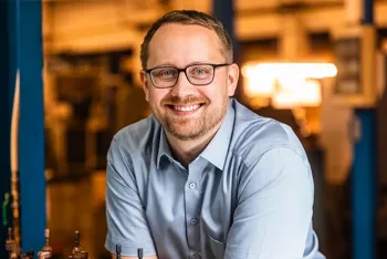 Ein lächelnder Mann mit Brille und gestutztem Bart, der ein hellblaues Button-up-Hemd trägt, steht in einer Industrie- oder Werkstattumgebung mit unscharfen Geräten im Hintergrund.