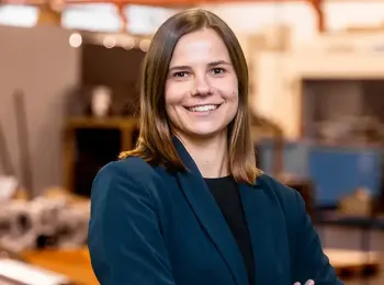 Eine Frau mit glattem braunem Haar, die einen dunklen Blazer und ein schwarzes Oberteil trägt, steht lächelnd mit verschränkten Armen in einem Innenraum oder einer industriellen Umgebung mit unscharfen Geräten im Hintergrund.