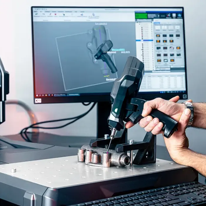 Eine Person scannt mit einem handgeführten 3D-Scanner ein Metallobjekt auf einem Tisch. Auf einem Computermonitor im Hintergrund wird eine 3D-Modellierungssoftware mit einem digitalen Rendering des gescannten Objekts angezeigt.