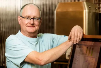 Ein Mann mit Brille und hellblauem Hemd lächelt, während er sich an einen Computermonitor lehnt, mit Industrieanlagen im Hintergrund.