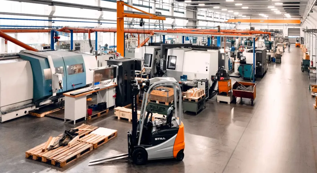 Eine geräumige, hell erleuchtete Fabrikhalle mit großen Industriemaschinen, Arbeitsplätzen und einem Gabelstapler. Paletten und Materialien sind im Raum verteilt, und große Fenster lassen natürliches Licht herein.