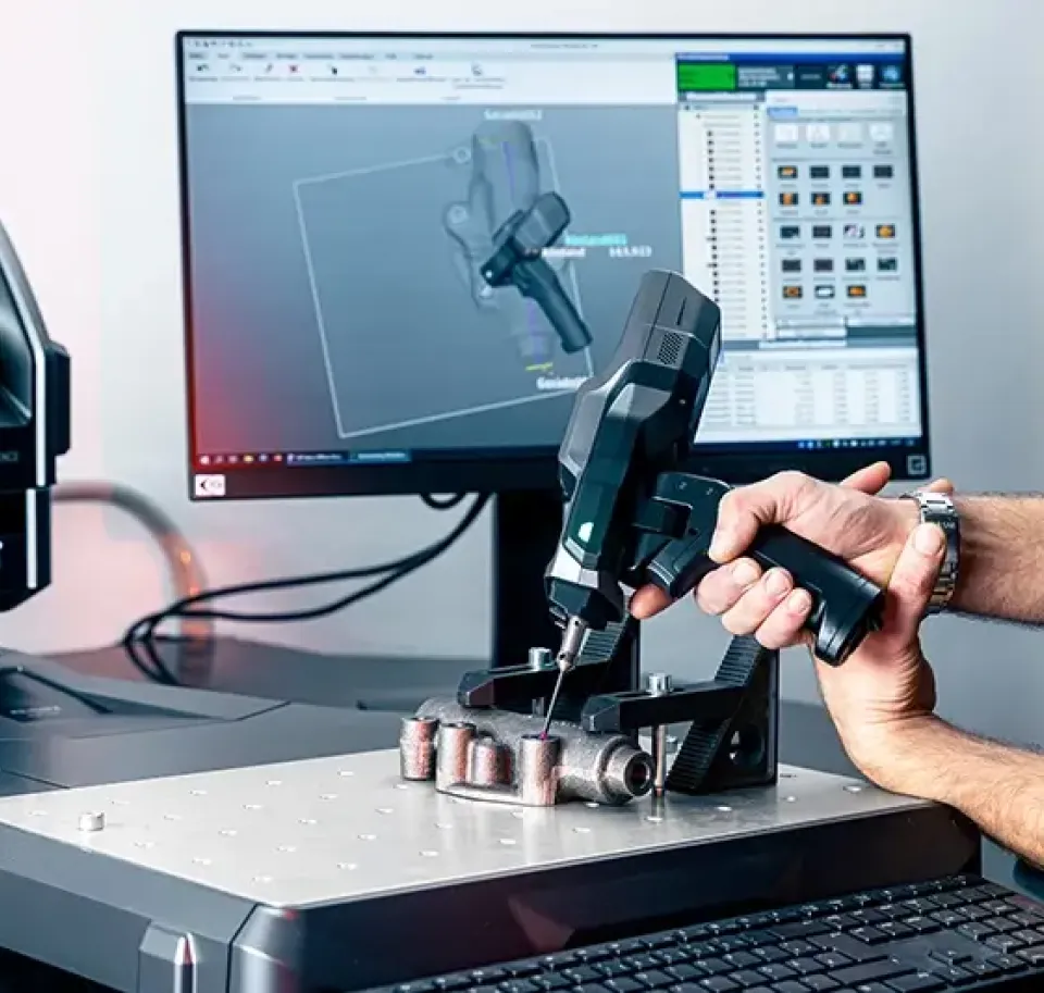 Eine Person scannt mit einem tragbaren 3D-Scanner ein Metallobjekt auf einer Plattform, während ein Computer im Hintergrund das 3D-Modell und die Scandaten anzeigt.