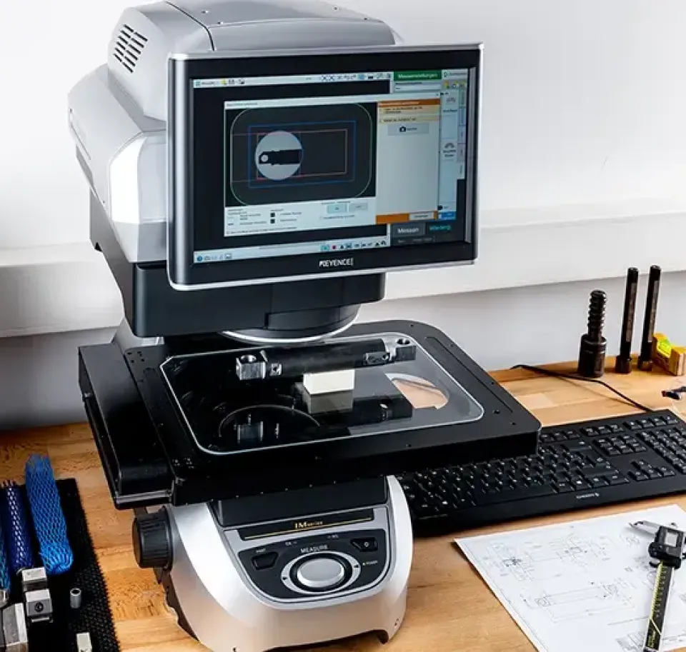 Ein digitaler optischer Komparator mit einem Monitor, einer Tastatur und verschiedenen Präzisionsmesswerkzeugen auf einem Holztisch, der für die Prüfung und Messung von Fertigungsteilen verwendet wird.