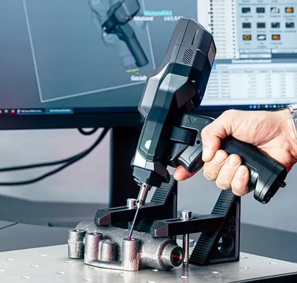 Eine Person scannt mit einem tragbaren 3D-Scanner ein Metallteil, wobei der Tastkopf des Scanners das Objekt berührt. Auf einem Computerbildschirm im Hintergrund wird eine 3D-Scansoftware angezeigt.