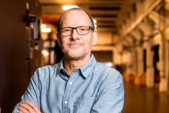 Ein Mann mittleren Alters mit kurzen Haaren, Brille und gestutztem Bart lächelt mit verschränkten Armen. Er trägt ein hellblaues Button-up-Hemd und steht in einem warm beleuchteten Industrieflur.