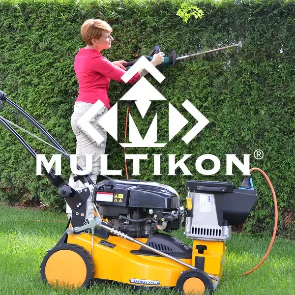 Eine Frau schneidet eine hohe grüne Hecke mit einer elektrischen Schere in einem Garten. Vor ihr steht eine gelbe Multikon-Rasenmaschine, auf der das Multikon-Logo und der Name gut sichtbar eingeblendet sind.